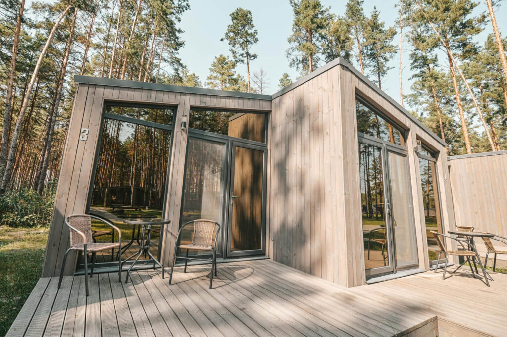 Modern wooden modular attached accessory dwelling unit with large glass panels and a front patio deck set among pine trees.